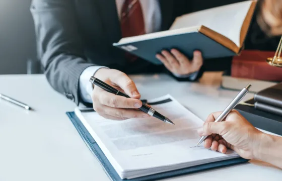 A Brief Overview Of Chapter 13 Bankruptcy Two people in business attire review and sign documents at a desk; one holds a book while the other signs a paper with a pen, consulting with a Cedar Rapids Chapter 7 bankruptcy attorney for financial guidance.