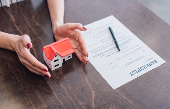 A Brief Overview Of Chapter 13 Bankruptcy A person places their hands around a small model house next to a bankruptcy form and a pen on a wooden table, symbolizing protection—an image often seen by a Cedar Rapids Chapter 7 bankruptcy attorney.